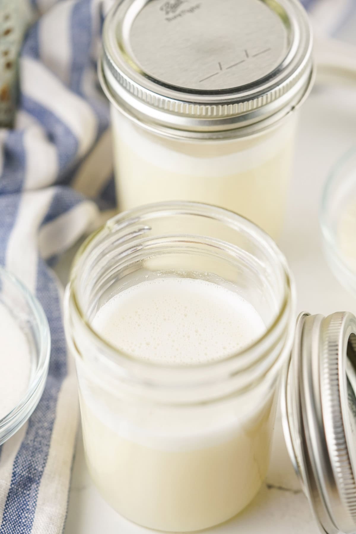 Two glass jars sit on a striped cloth, one open and filled with creamy homemade sweetened condensed milk, the other closed with a metal lid. The scene suggests a freshly prepared dairy treat ready to enjoy.