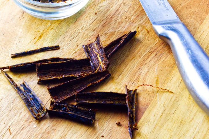 Sliced vanilla bean pods on a wooden cutting board next to a metal knife, with a glass bowl partially visible in the corner.