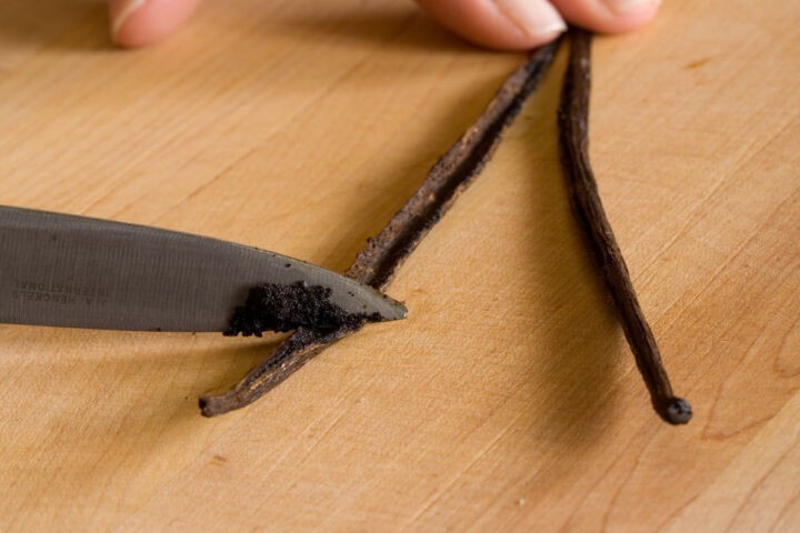 A knife is scraping vanilla seeds from an open vanilla bean pod on a wooden surface, with a hand holding the pod steady.