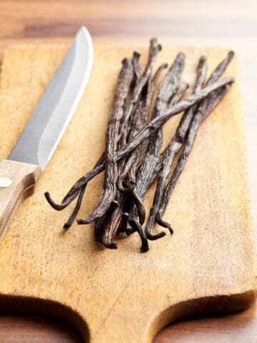 A pile of dried vanilla beans lies on a wooden cutting board next to a kitchen knife with a wooden handle, all set on a wooden surface.