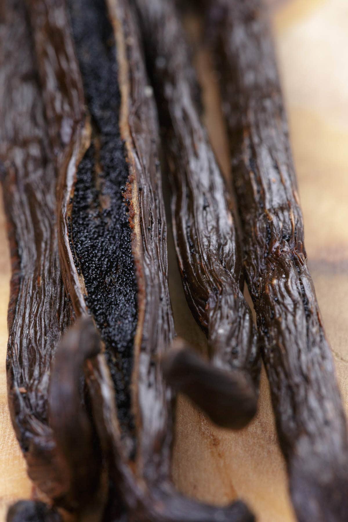 Close-up of dried vanilla beans on a wooden surface, with one pod split open to reveal tiny black vanilla seeds inside.