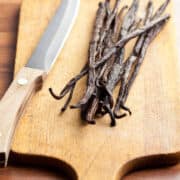 A wooden cutting board with a few whole dried vanilla beans and a kitchen knife with a wooden handle placed on top.