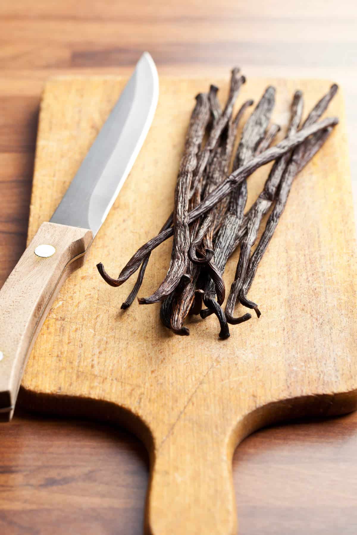 A wooden cutting board with a few whole dried vanilla beans and a kitchen knife with a wooden handle placed on top.