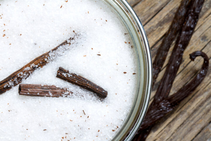 A glass jar filled with white granulated sugar and pieces of vanilla bean, placed on a wooden surface. Two whole vanilla beans lie next to the jar.