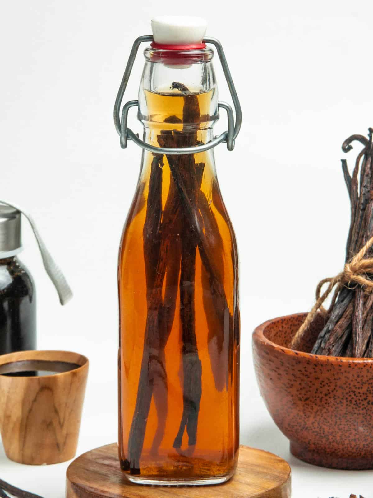 A clear glass bottle filled with amber liquid and vanilla beans, sealed with a white cap and metal clasp, sits on a wooden coaster. Nearby are a wooden bowl of vanilla beans, a small wooden cup, and a dark bottle.