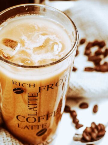 A glass of iced cafe au lait with milk rests on a textured cloth, surrounded by scattered coffee beans. The glass features decorative text, including words like "latte" and "coffee.