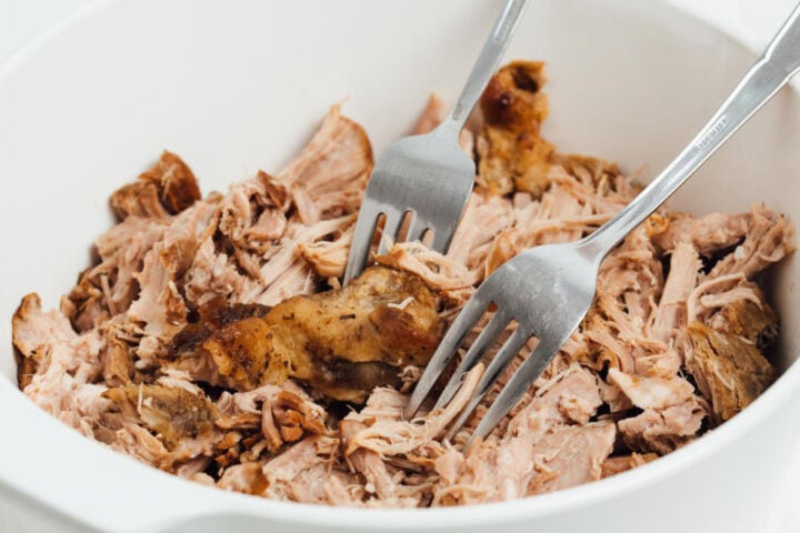 Two forks pulling apart tender cooked pork butt in a white dish.