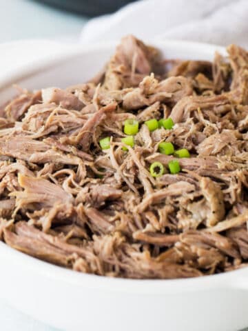 Tender Instant Pot pork butt shredded in a white serving dish with tongs nearby.