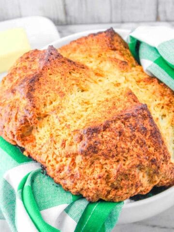 A golden-brown loaf of Irish soda bread sits in a bowl lined with a green and white checkered cloth, with a stick of butter in the background on a marble surface-perfect for enjoying authentic Irish soda bread at home.