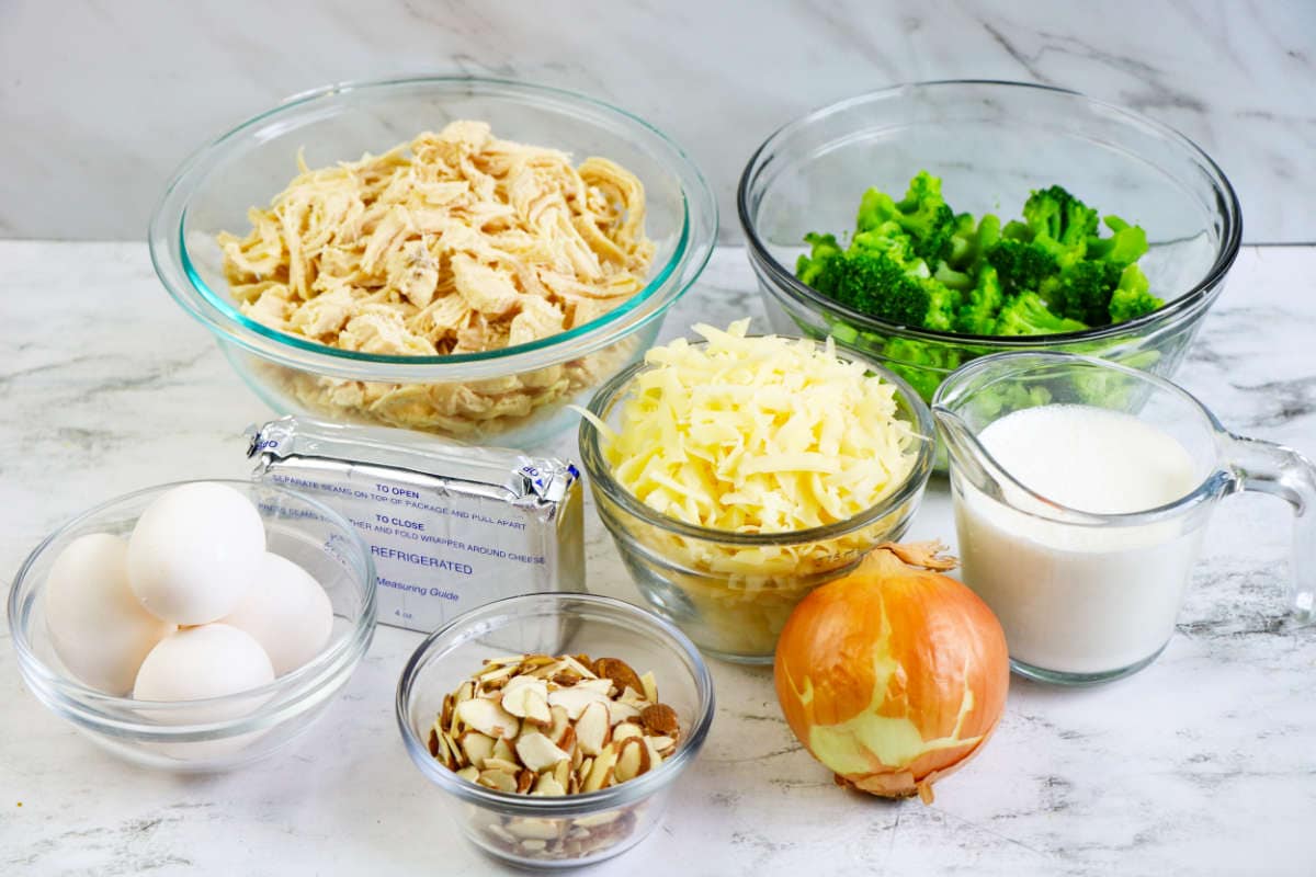 A variety of ingredients on a white surface, perfect for a Low Carb Chicken and Swiss casserole, including shredded chicken, broccoli, shredded cheese, sliced almonds, an onion, eggs, cream cheese, and a glass measuring cup of milk.