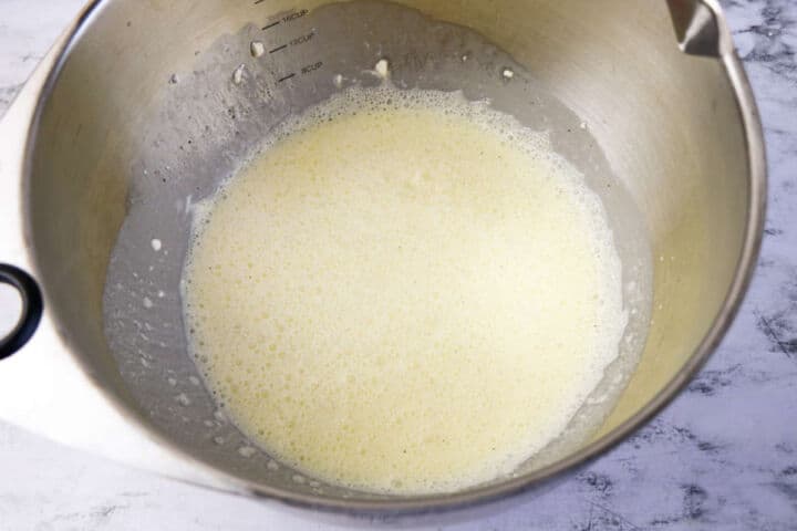 A stainless steel mixing bowl contains a pale, frothy liquid mixture, ready for making Low Carb Chicken and Swiss casserole, sitting on a light marble countertop. Measurement markings are visible on the inside of the bowl.