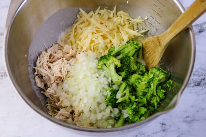 A metal mixing bowl holds ingredients for a Low Carb Chicken and Swiss casserole, with shredded chicken, chopped onions, broccoli florets, and shredded cheese. A wooden spoon rests on the side of the bowl.
