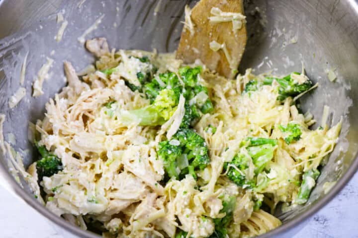 A metal mixing bowl filled with shredded chicken, broccoli florets, shredded cheese, and a creamy sauce is being stirred with a wooden spoon-perfect as the base for a Low Carb Chicken and Swiss casserole.
