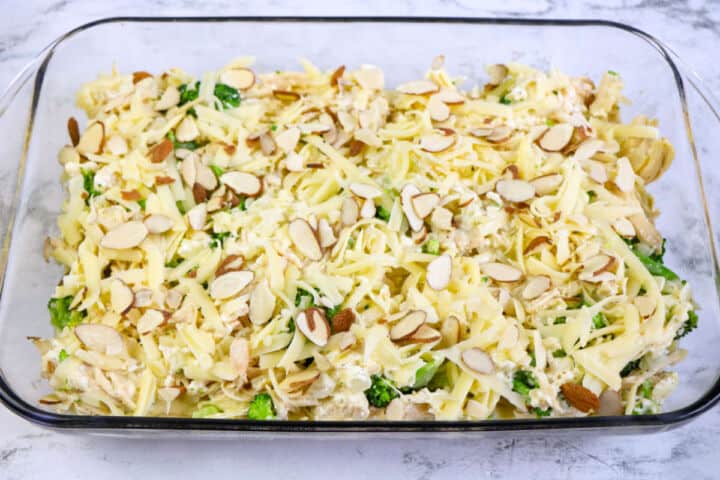 A glass baking dish filled with Low Carb Chicken and Swiss casserole sits on a white marble surface, topped with shredded cheese and sliced almonds. Pieces of broccoli and bits of chicken are visible under the cheese.