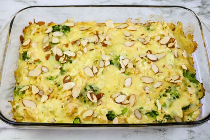 A Low Carb Chicken and Swiss casserole in a glass dish, topped with melted cheese, sliced almonds, and visible broccoli pieces sits on a light-colored countertop.