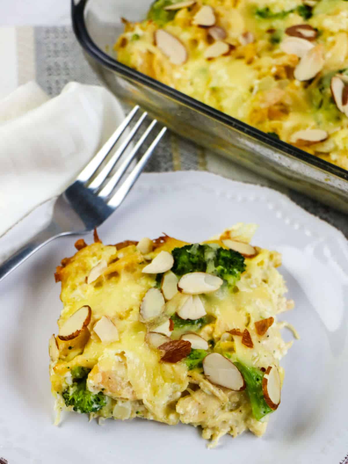 A slice of cheesy Low Carb Chicken and Swiss casserole topped with sliced almonds sits on a white plate next to a fork and napkin, with the rest of the casserole in a baking dish in the background.