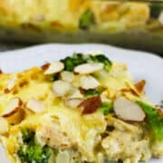 A slice of Low Carb Chicken and Swiss casserole with broccoli, topped with sliced almonds on a white plate, while the remaining cheesy casserole rests in a glass baking dish in the background.