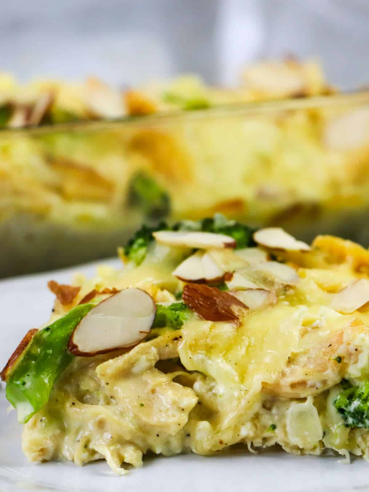 A close-up of a cheesy Low Carb Chicken and Swiss casserole slice, topped with sliced almonds, on a white plate, with the casserole dish blurred in the background.