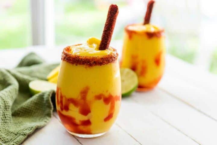 Two Mangonada smoothies in clear glasses, swirled with red sauce, garnished with chili powder on the rim and a tamarind candy stick. A green cloth and lime halves are nearby on a white surface.