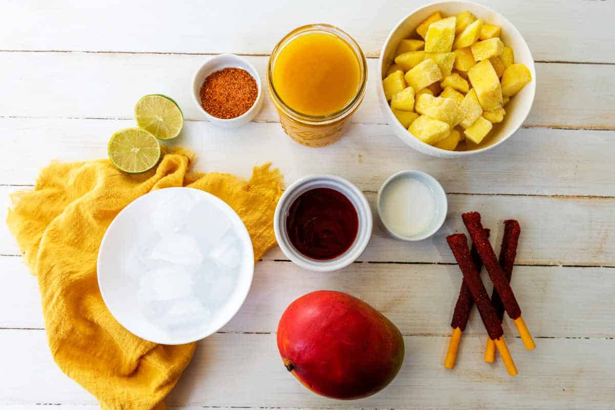 Assorted ingredients on a white table for a classic chamoyada: a mango, frozen mango chunks, chamoy sauce, lime, jar of mango juice, small bowls of tajin and milk, ice cubes, and three tamarind candy sticks for the perfect Mangonada treat.
