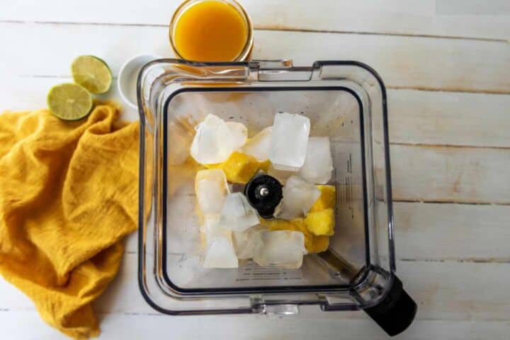 A blender containing ice cubes and pineapple chunks sits on a white wooden surface, ready for a tropical twist on a Mangonada. Nearby are a glass of orange juice, two lime halves, and a yellow cloth.