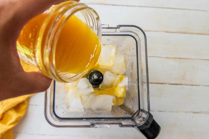 A hand pours orange juice from a glass jar into a blender filled with ice cubes, creating the perfect base for a refreshing Mangonada, viewed from above on a light wooden surface.