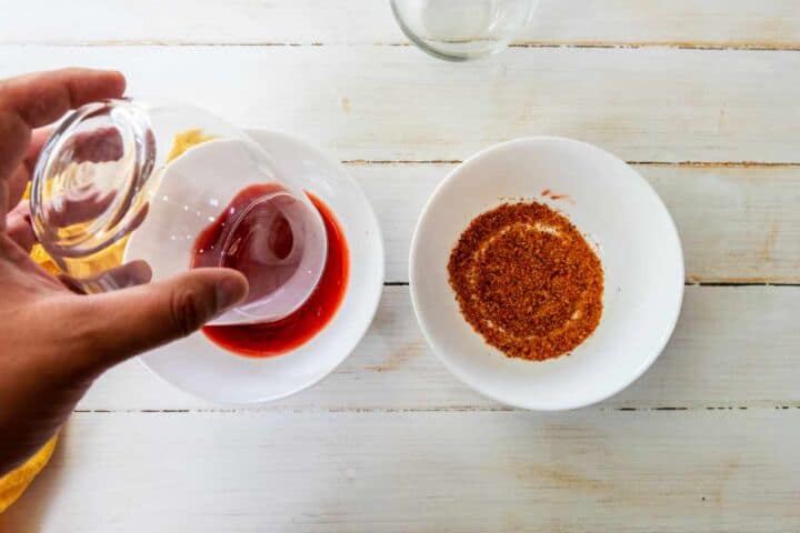 A hand dips the rim of a glass into red liquid on a white plate, beside reddish-brown seasoning—perfect prep for a homemade Mangonada on a white wooden surface.