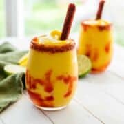A glass of bright yellow Mangonada smoothie with red chamoy sauce swirls, topped with a tamarind candy stick and chili-lime seasoning on the rim, with another chamoyada, lime slices, and a green napkin in the background.