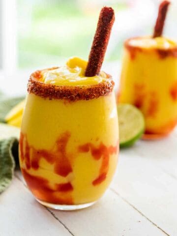 A glass of bright yellow Mangonada smoothie with red chamoy sauce swirls, topped with a tamarind candy stick and chili-lime seasoning on the rim, with another chamoyada, lime slices, and a green napkin in the background.