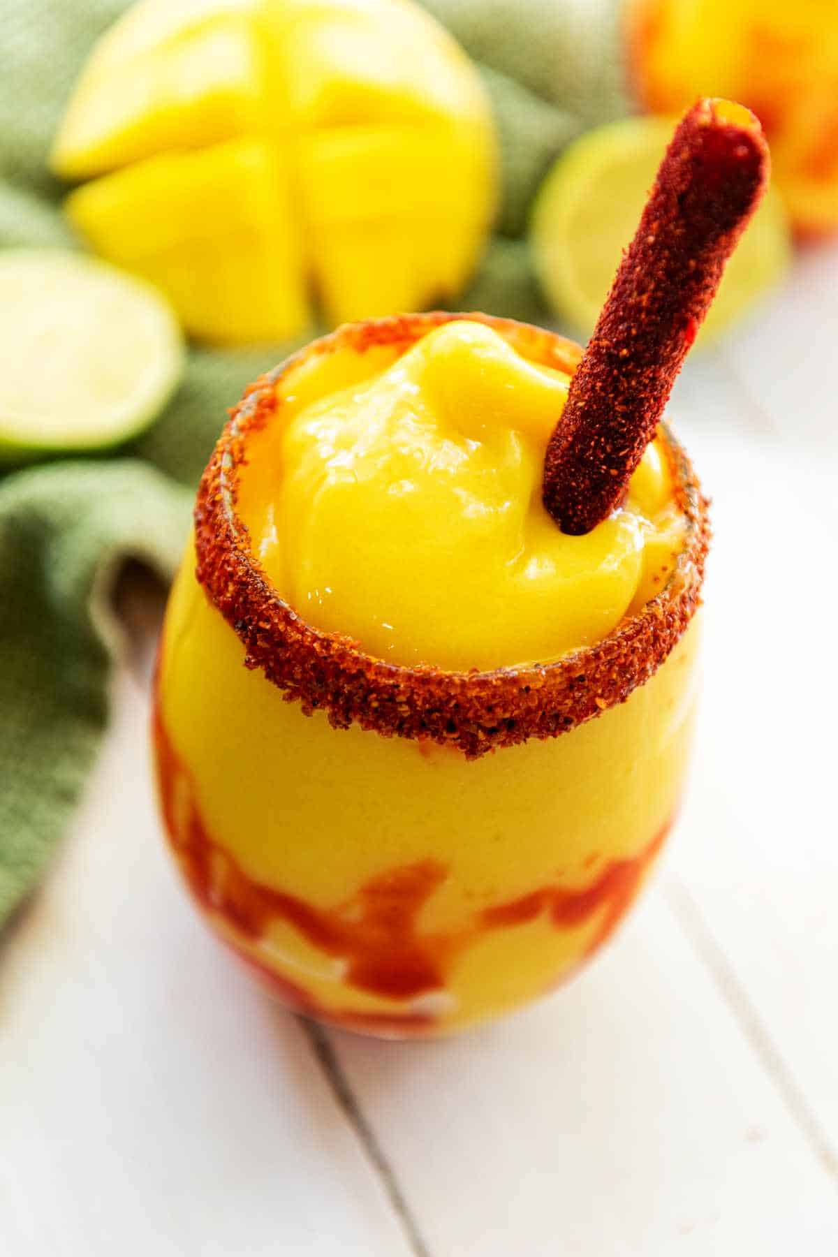 A glass of bright yellow mangonada smoothie with a spicy chili powder rim and a tamarind candy stick, chamoy sauce drizzled inside the glass. Sliced mango and limes are blurred in the background.