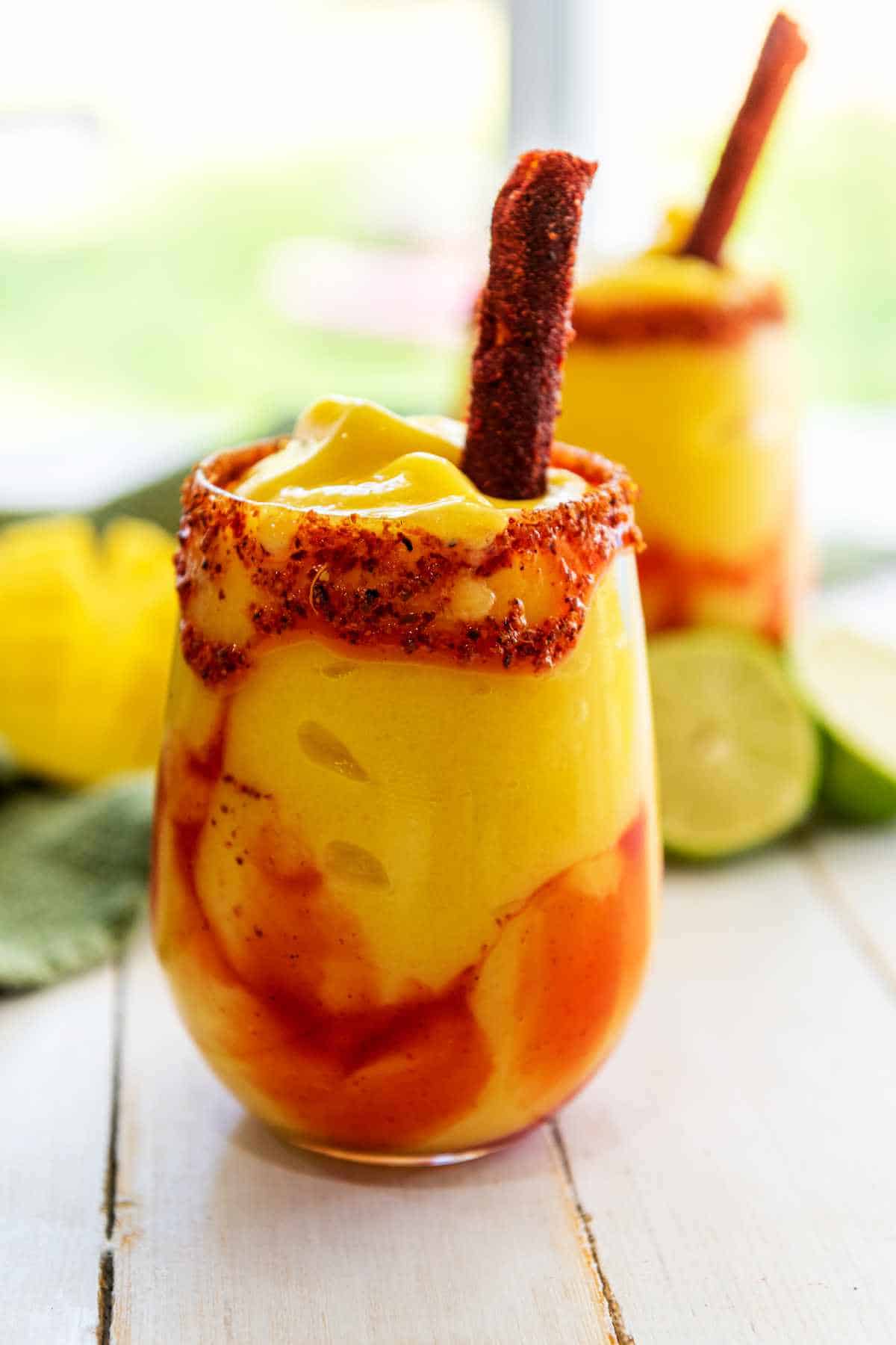 A glass of mango chamoyada, or mangonada, topped with chili powder, chamoy sauce, and a tamarind candy stick, with a second glass, lime, and yellow fruit blurred in the background.