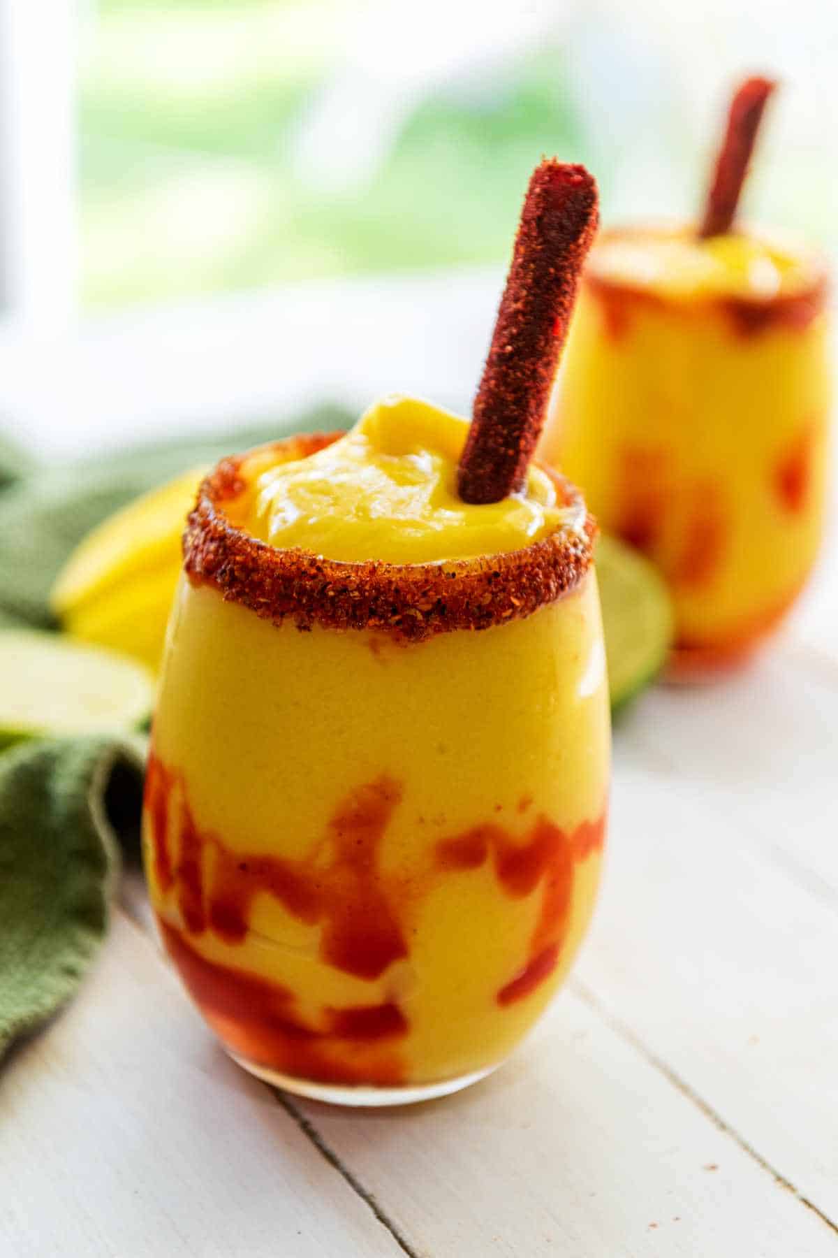 A glass of bright yellow mangonada smoothie with swirls of red chamoy sauce, a chili powder rim, and a tamarind candy stick. Another similar chamoyada drink is blurred in the background on a white surface.
