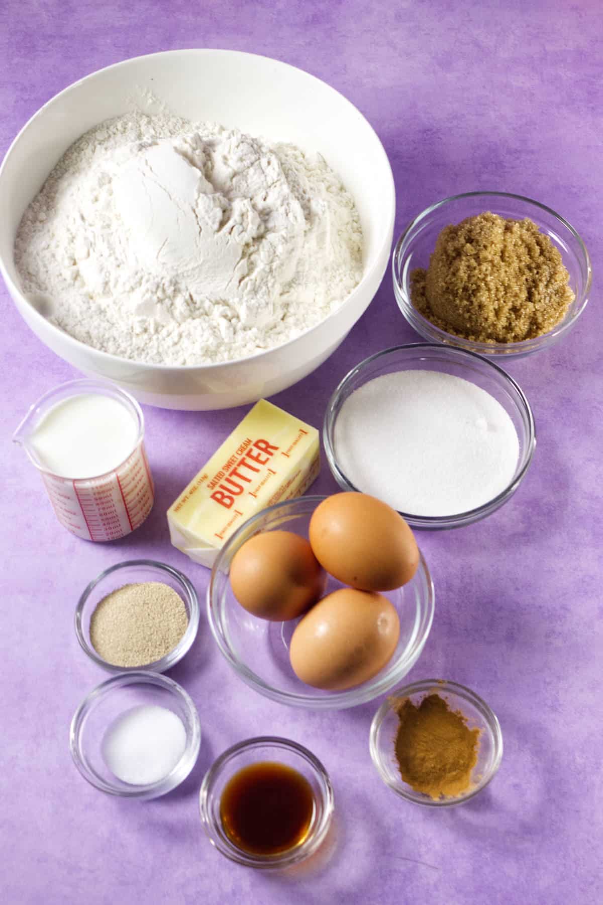 A variety of baking ingredients on a purple surface, perfect for making mardi gras king cake, including a bowl of flour, brown sugar, white sugar, three eggs, a stick of butter, milk, yeast, salt, vanilla, and ground cinnamon.