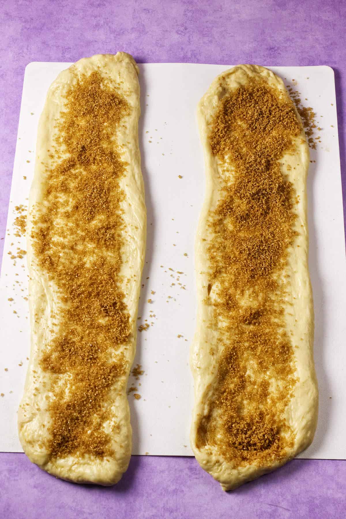 Two long strips of raw dough, ready to become Mardi Gras king cake, are topped with brown sugar and sit side by side on a white surface against a purple background.