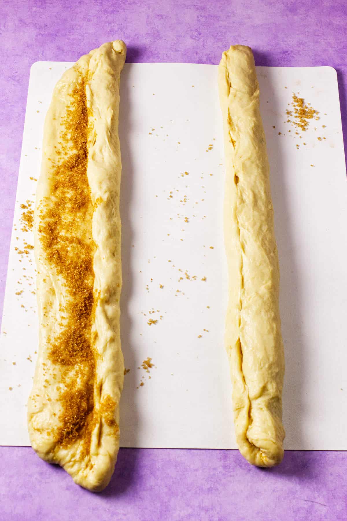 Two strips of dough on a white surface, one sprinkled with brown sugar filling and the other rolled up, ready for baking a mardi gras king cake, all set against a vibrant purple background.
