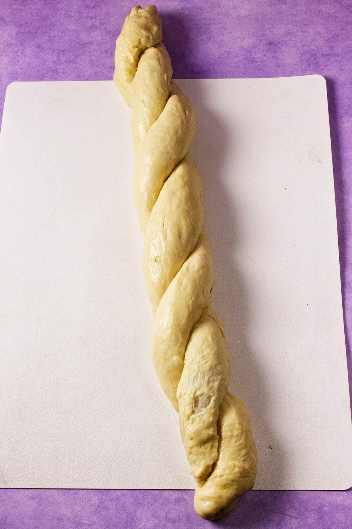A thick, braided strand of unbaked dough-perfect for a Mardi Gras king cake-rests on a white cutting board with a purple background.