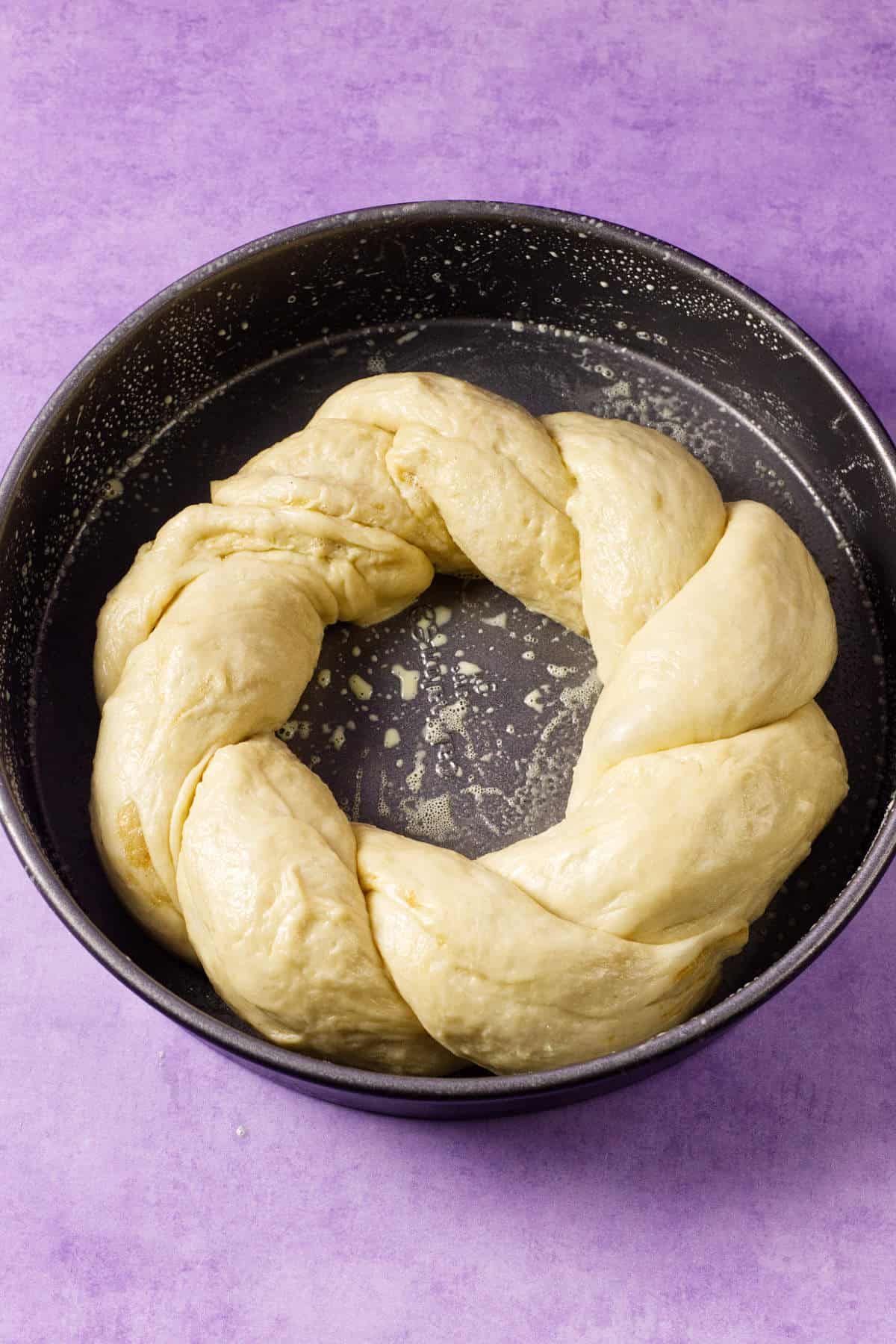 A round baking pan with unbaked braided dough arranged in a ring shape, ready to become a classic Mardi Gras king cake, set on a light purple surface.