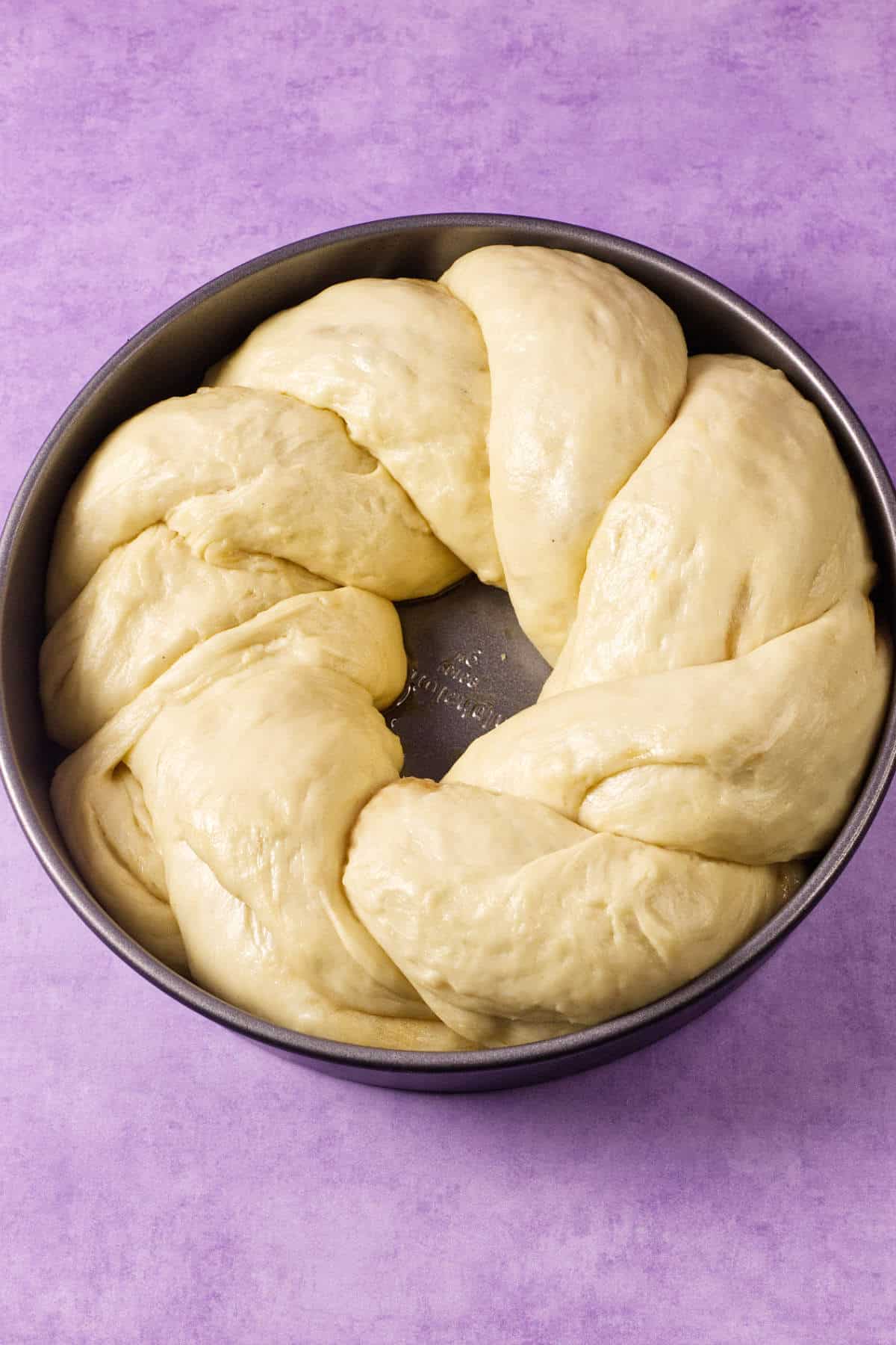 A round baking pan with braided, unbaked dough arranged in a circle, ready to become a festive Mardi Gras king cake, rests on a light purple surface.
