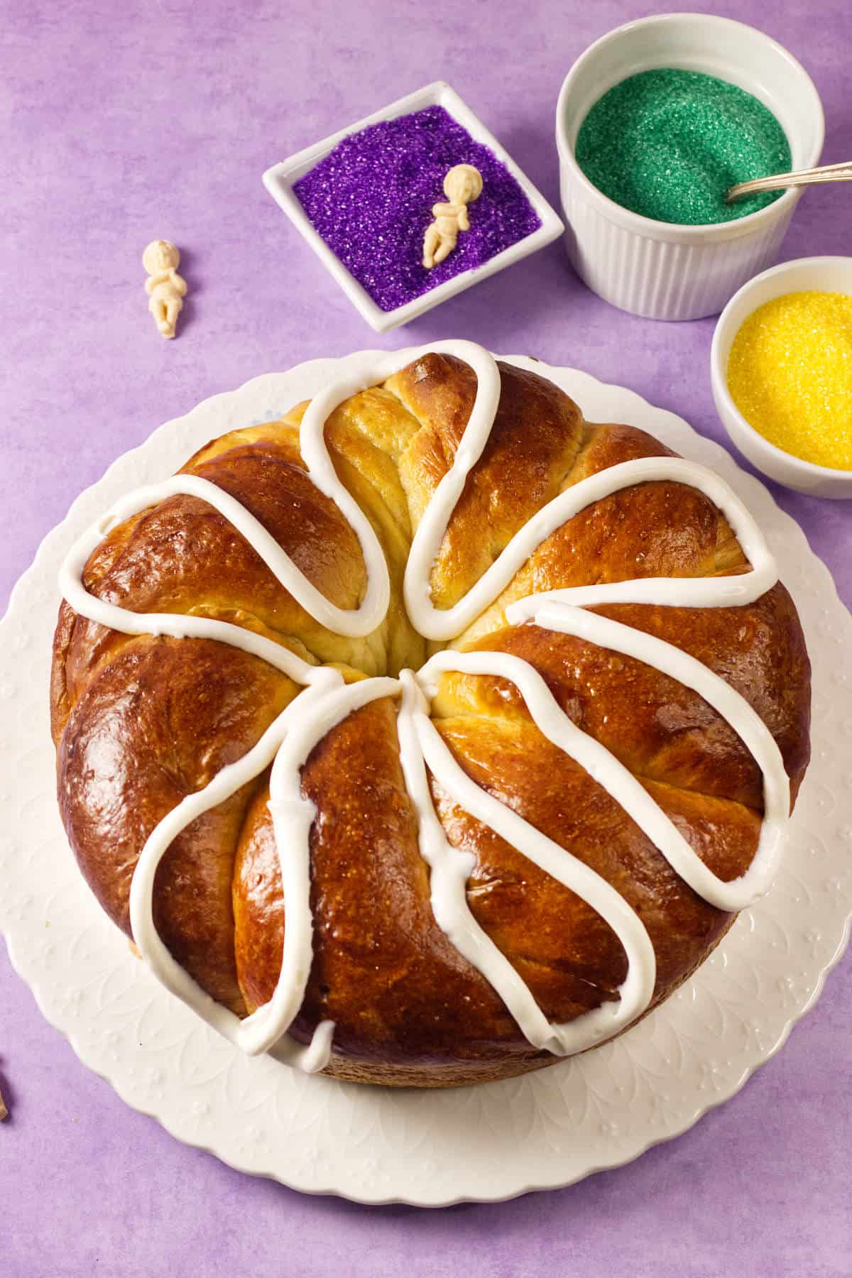 A round Mardi Gras King Cake with a shiny brown crust and white icing sits on a white plate. Bowls of purple, yellow, and green sugar sprinkles rest nearby on a purple surface.