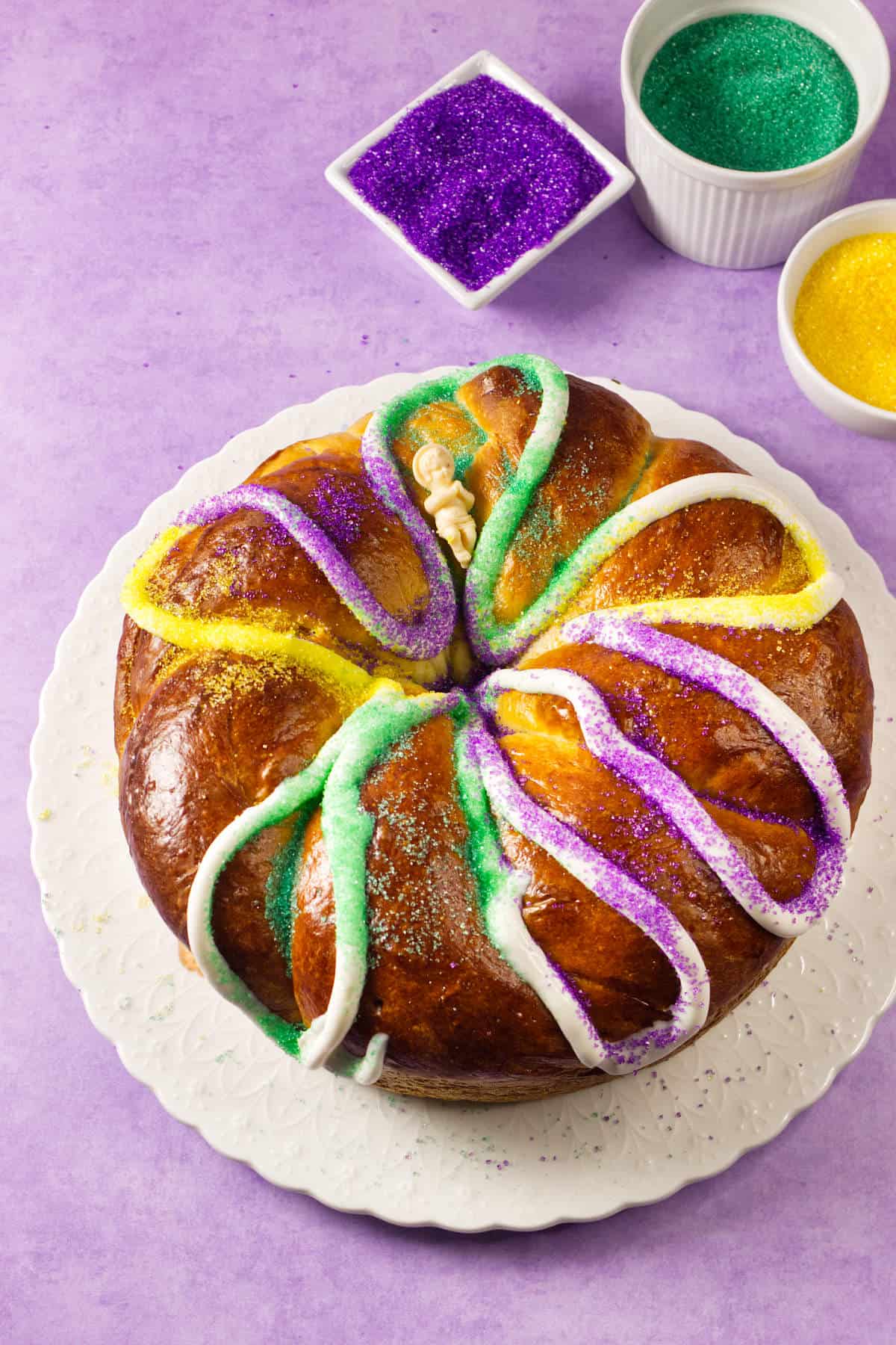A round Mardi Gras King Cake decorated with white icing and sprinkled with yellow, green, and purple sugar sits on a white plate. Small bowls of colored sugar are nearby on a vibrant purple background.