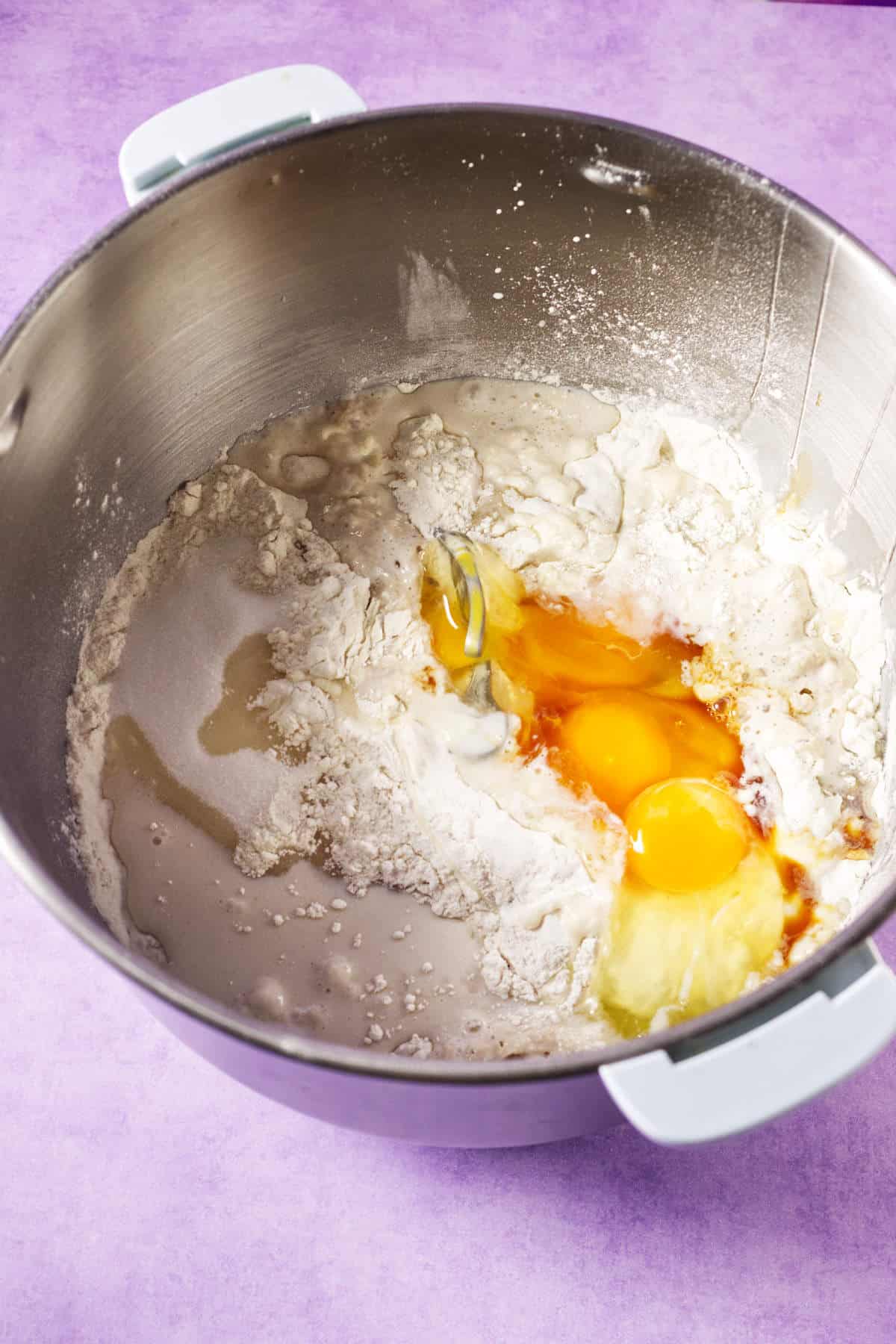 A metal mixing bowl containing flour, two cracked eggs, vanilla extract, and other liquid ingredients for a Mardi Gras king cake sits ready to be mixed on a purple surface.