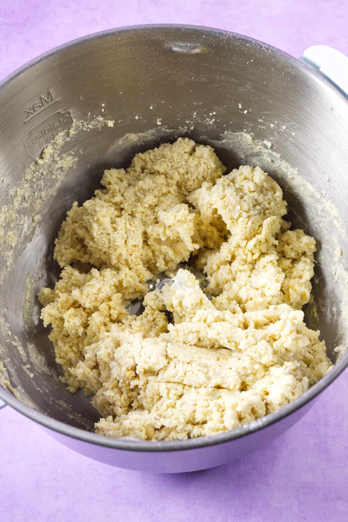 A large metal mixing bowl filled with pale, crumbly cookie dough sits on a light purple surface, resembling the festive beginnings of a Mardi Gras king cake. The dough appears freshly mixed and is slightly uneven in texture.