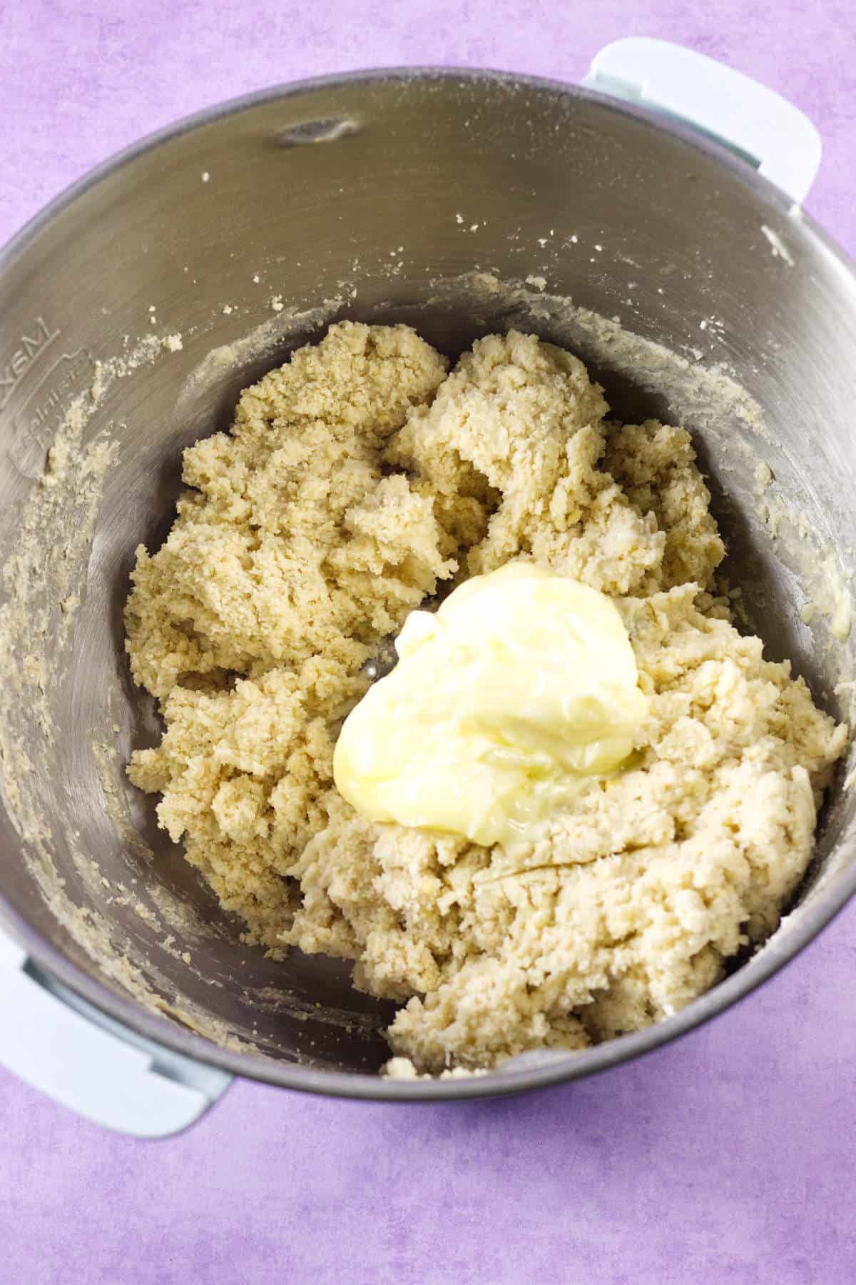 A mixing bowl containing partially mixed cookie dough with a dollop of butter on top, reminiscent of preparing a Mardi Gras king cake, all set against a purple background.