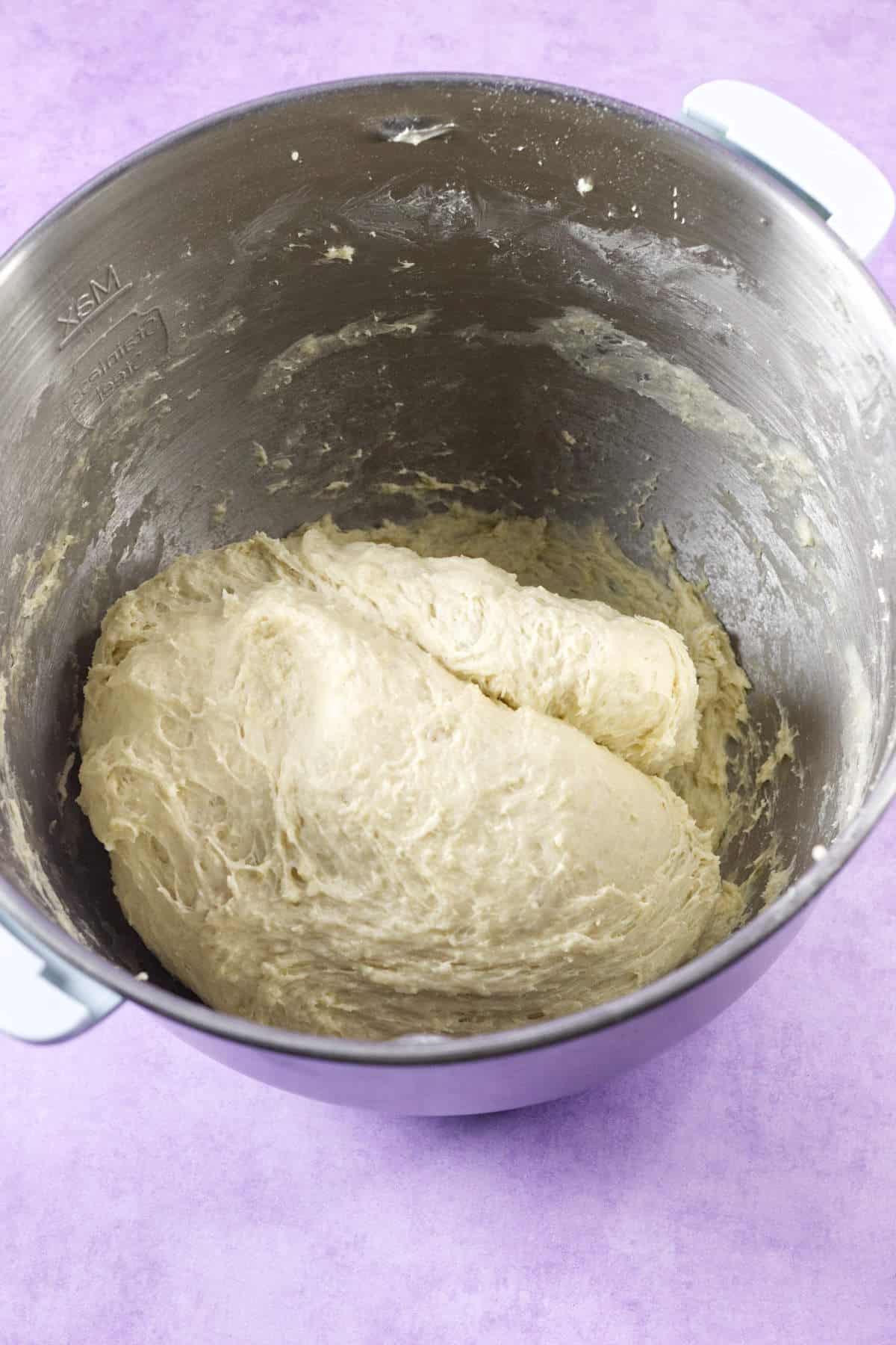 A stainless steel mixing bowl filled with sticky, unbaked mardi gras king cake dough sits on a light purple surface.