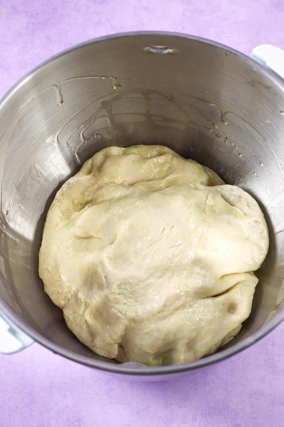 A ball of raw dough for mardi gras king cake sits in a large stainless steel mixing bowl, with the bowl resting on a light purple surface.
