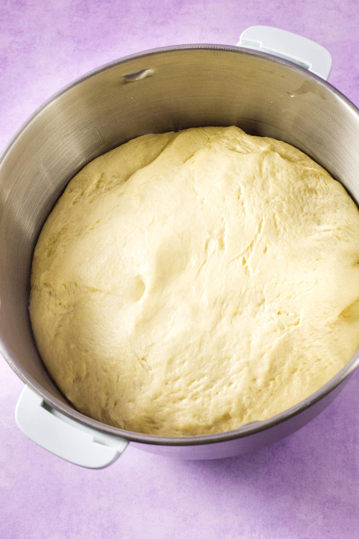 A large metal mixing bowl filled with risen, fluffy mardi gras king cake dough sits on a light purple surface.
