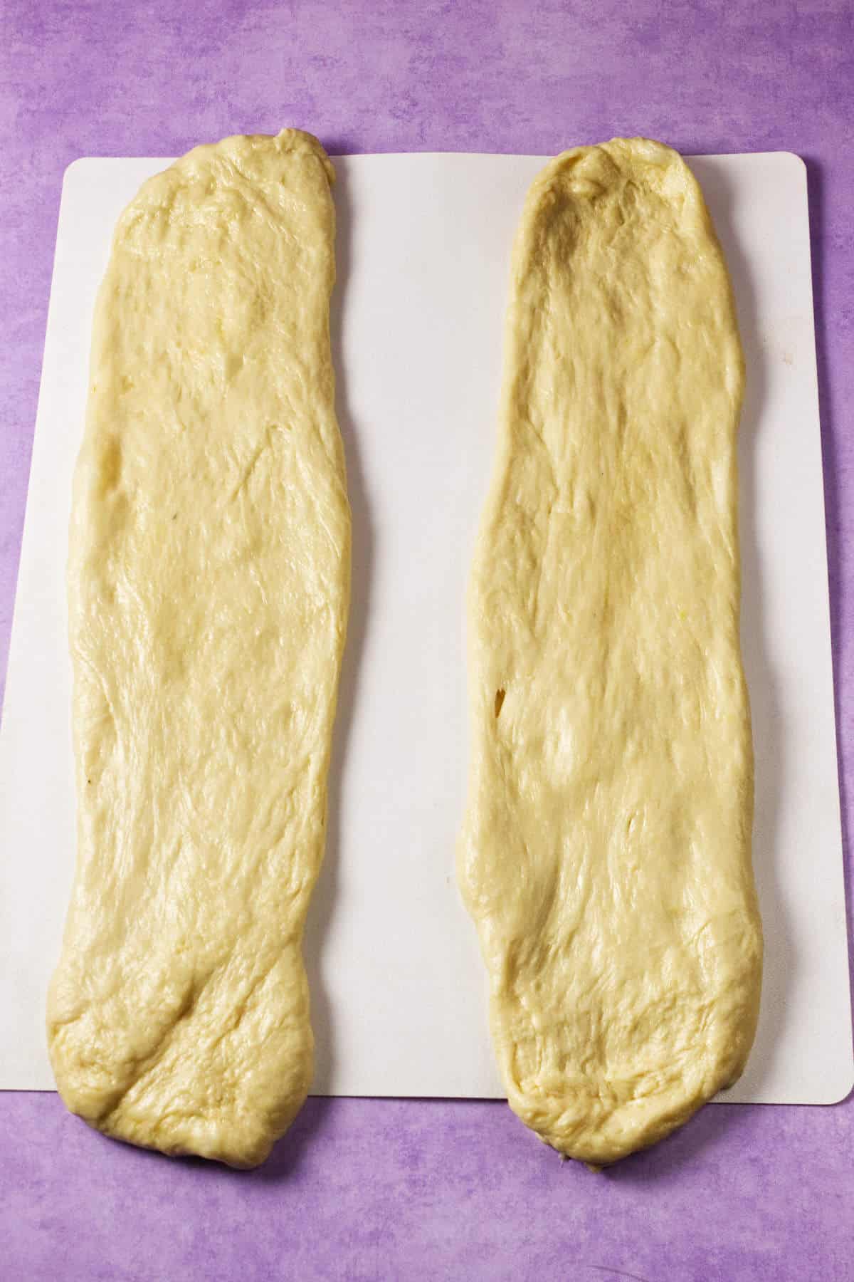 Two stretched, rectangular pieces of dough-perfect for making a Mardi Gras king cake-are placed side by side on a white cutting board, set on a purple surface.