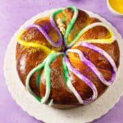 A round King Cake, a festive Mardi Gras treat, is decorated with white icing and sprinkled with green, yellow, and purple sugar. It sits on a white plate atop a purple surface.