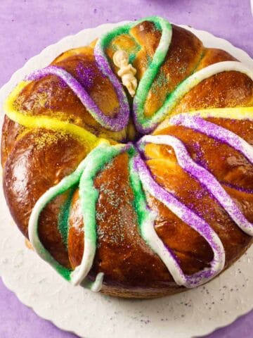 A round King Cake, a festive Mardi Gras treat, is decorated with white icing and sprinkled with green, yellow, and purple sugar. It sits on a white plate atop a purple surface.
