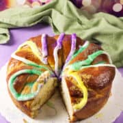 A round Mardi Gras king cake decorated with colorful purple, yellow, and green sugar and icing sits on a white plate with a slice cut out. A green cloth and festive blurred lights are in the background.
